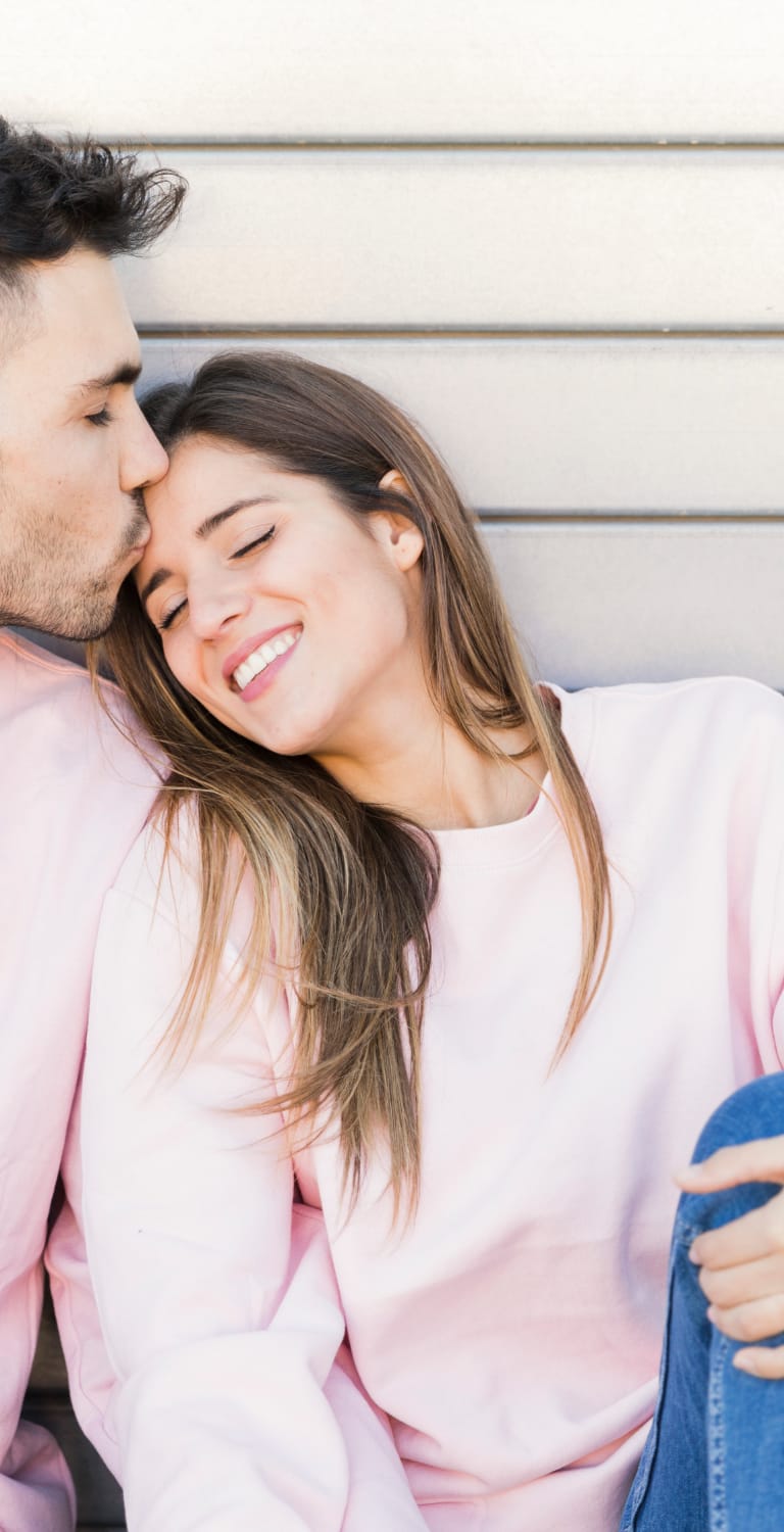 Couple with matching pink sweaters sitting on a bench with man kissing womans forehead